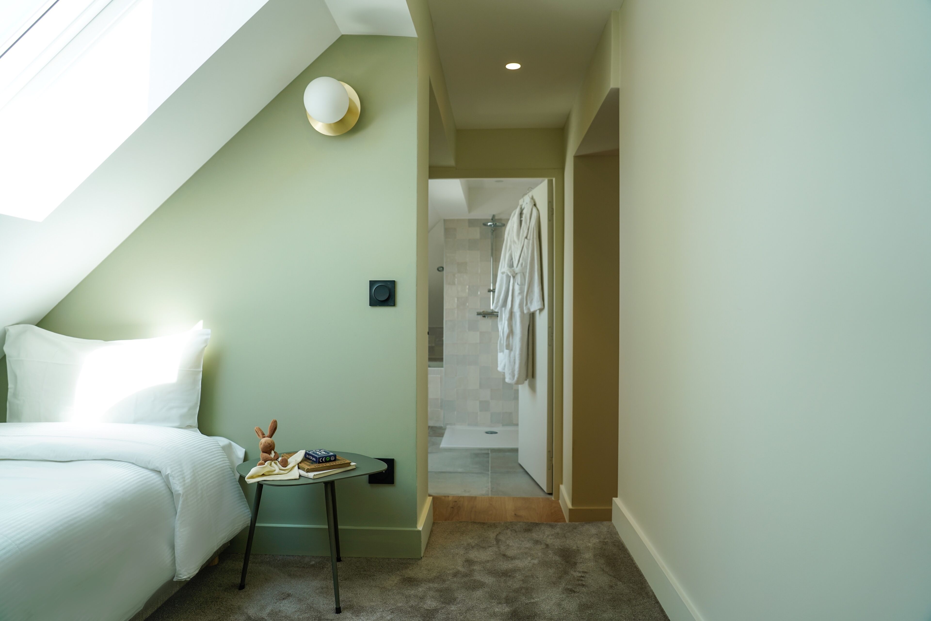 A bedroom with a bed, a small table with a book and a toy, and a skylight.
