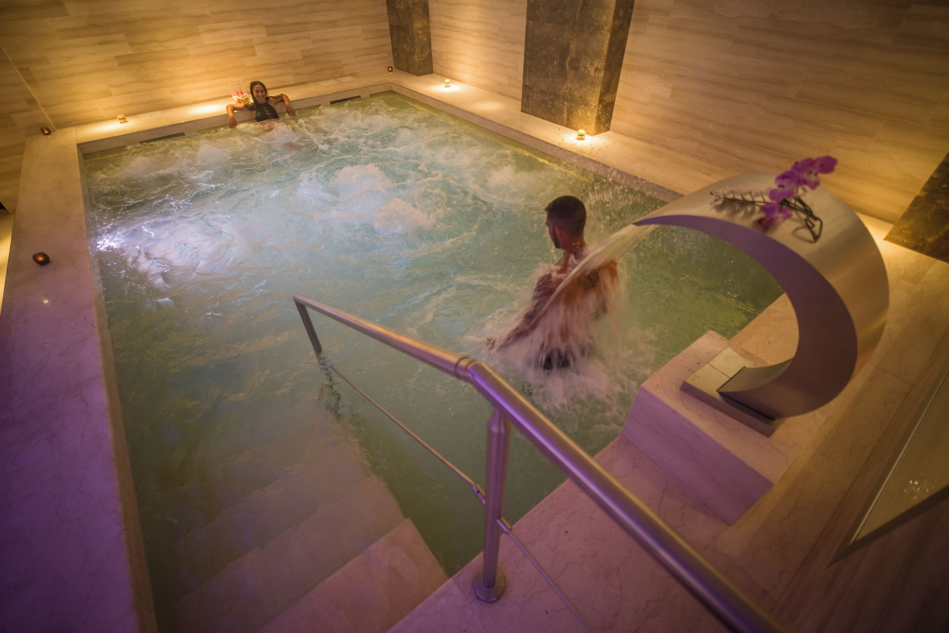 Une personne dans un grand jacuzzi intérieur, avec une rambarde en métal et une composition florale décorative.