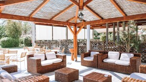 An outdoor patio area with wicker furniture, including sofas and ottomans, under a wooden pergola with a thatched roof.