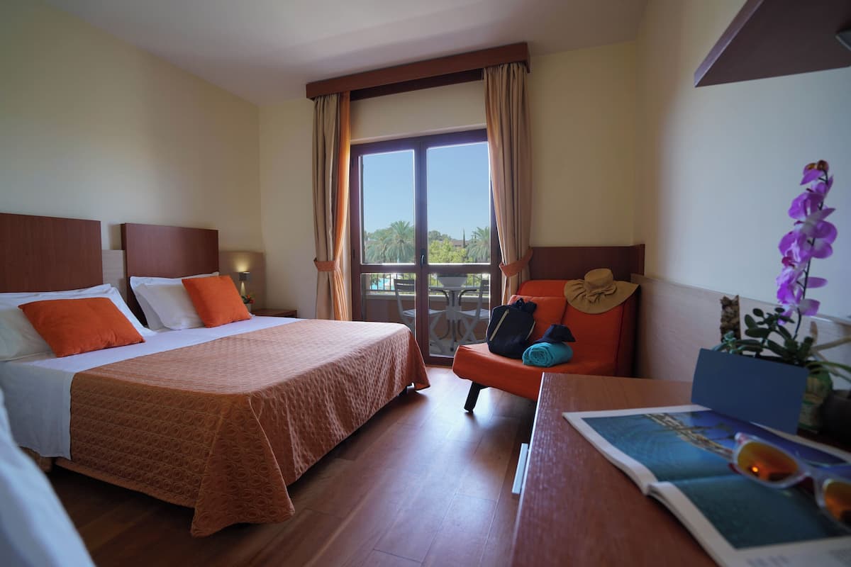 Une chambre d’hôtel avec deux lits, un balcon, un bureau avec une chaise et une fenêtre avec des rideaux.