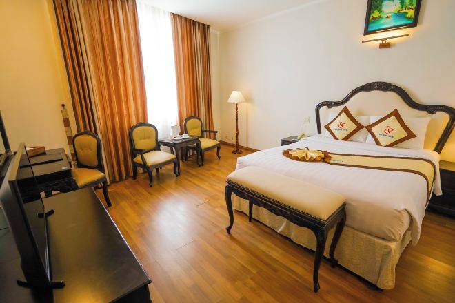 A hotel room with a bed, a desk, two chairs, a lamp, and a painting on the wall.