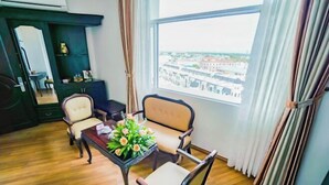 Quarto com piso de madeira, uma mesa com um vaso de flores, duas cadeiras, um armário escuro e uma janela com vista para edifícios.