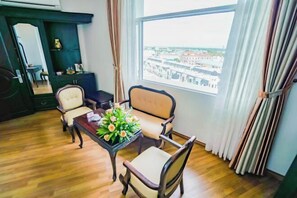 Una habitación con piso de madera, una mesa con un florero, dos sillas, un mueble oscuro y un ventanal con vistas a edificios.