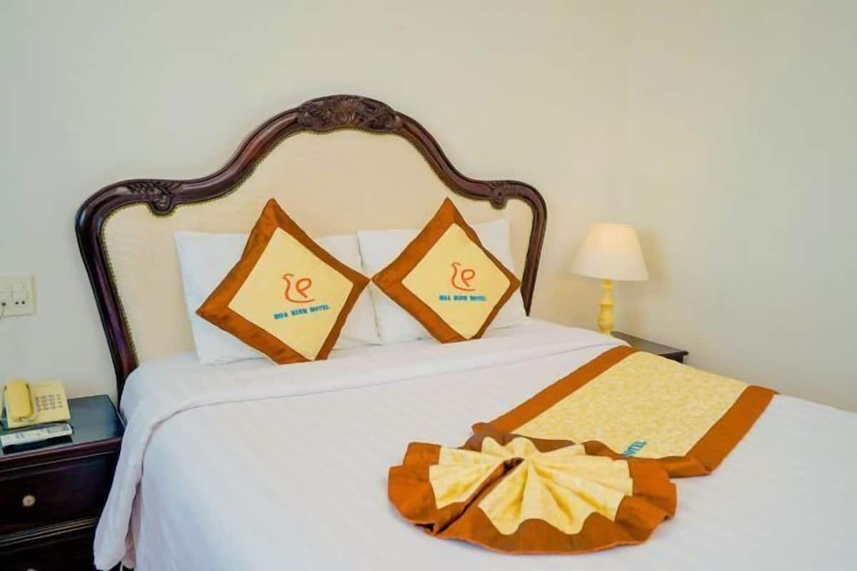Una cama bien hecha con un cojín decorativo, una lámpara de noche y un teléfono en la mesita.