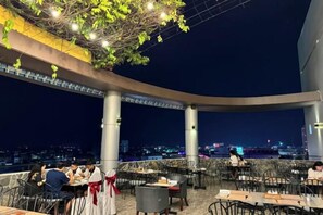 Area pranzo all'aperto con tavoli e sedie, vista sulla città e una zona coperta con luci a sospensione.