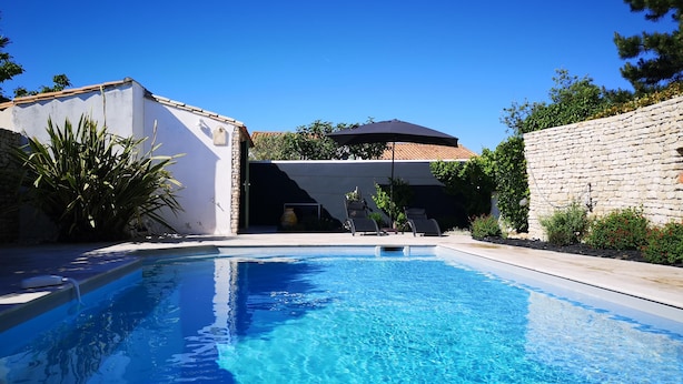 Ein Schwimmbecken mit klarem, blauem Wasser, umgeben von einer Steinmauer und einem weißen Gebäude mit einem Ziegeldach.