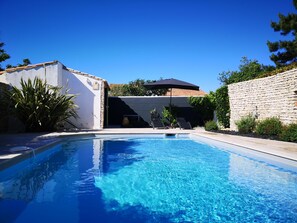 Una piscina con agua azul cristalina, rodeada por un muro de piedra y un edificio blanco con tejado de tejas.