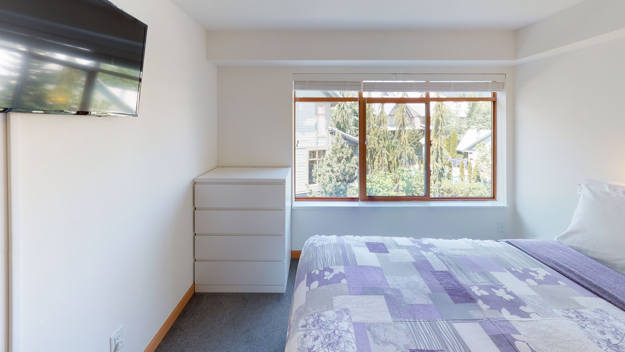 A bedroom with a bed, a window, a dresser, and a wall-mounted TV.