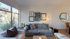 Una sala de estar moderna con un sofá gris, un sillón blanco y una mesa de centro. Hay obras de arte enmarcadas en la pared, una lámpara de pie y un espejo redondo.