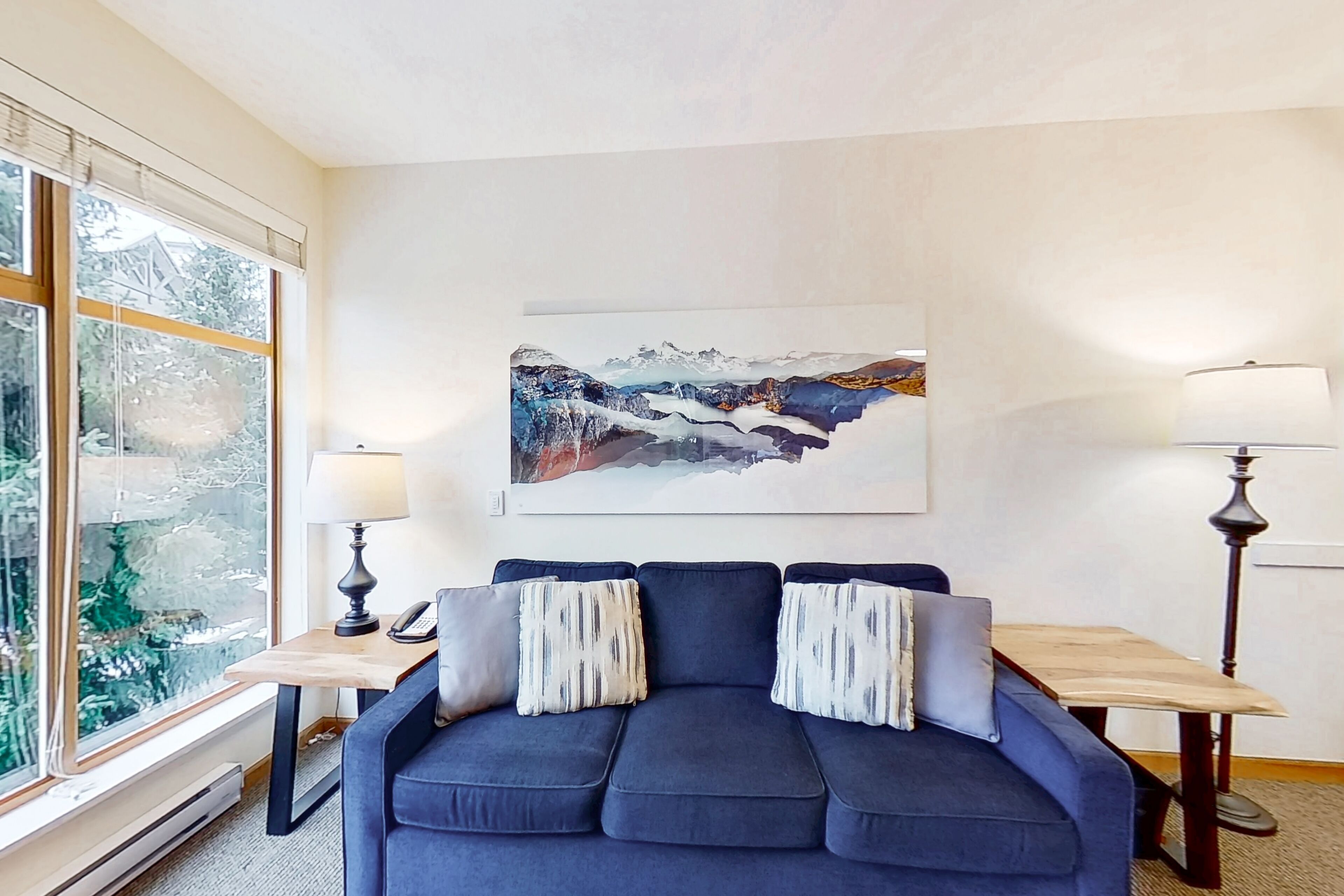 Un salon avec un canapé bleu foncé, deux tables d’appoint avec des lampes, un grand tableau abstrait accroché au mur et une fenêtre à vitrage dépoli.