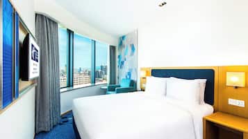 A hotel room with a large bed, a TV mounted on the wall, a city view through the window, and a small table with a lamp.