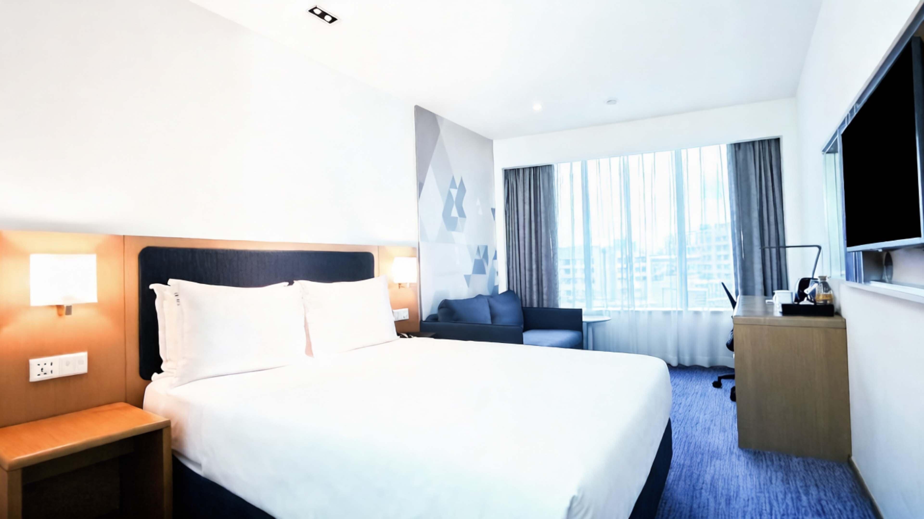 A hotel room with a large bed, a desk with a lamp, a sofa, and a TV on the wall.
