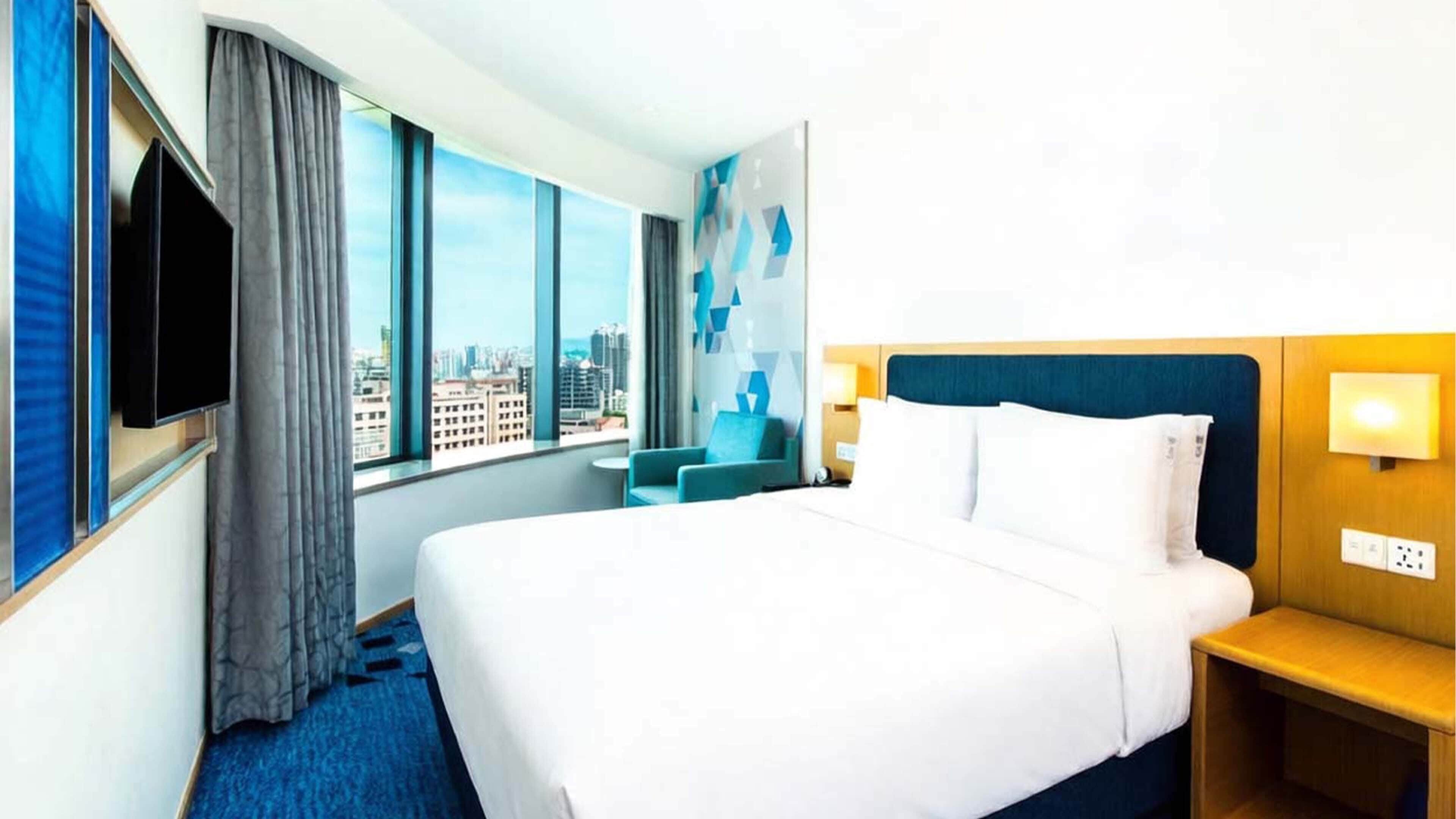 A hotel room with a large bed, a TV mounted on the wall, a seating area with a chair, and a view of the city through large windows.