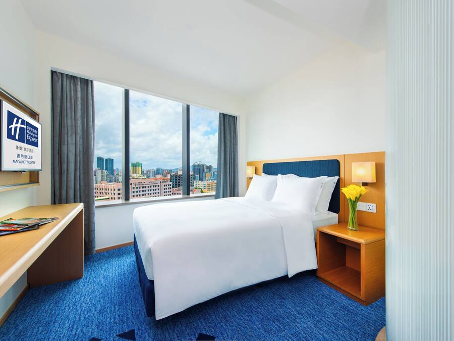Quarto de hotel com cama, escrivaninha, televisão e janela com vista para a cidade.