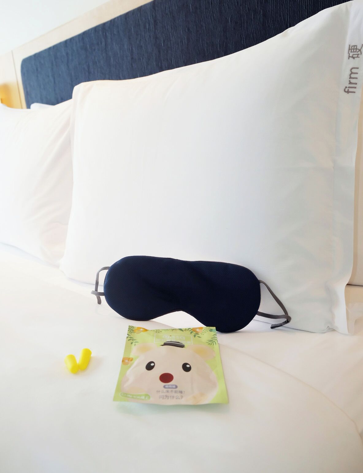 Una cama con ropa de cama blanca, una almohada azul oscuro, una antifaz azul oscuro y una pequeña bolsita de tapones para los oídos.