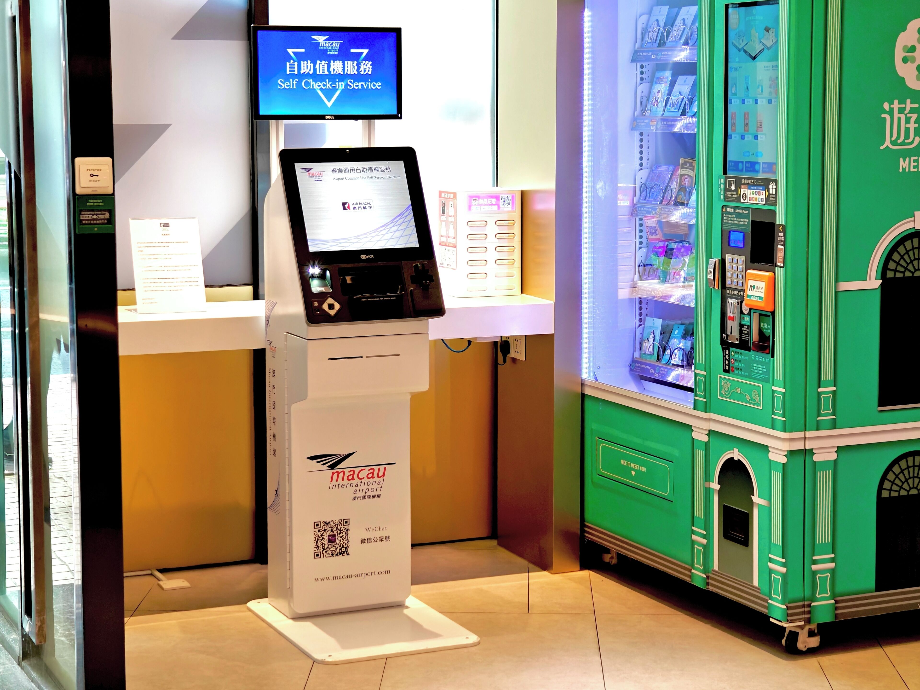 Quiosco de autoservicio en un aeropuerto con pantalla digital, un código QR y un cartel para el Aeropuerto Internacional de Macao.