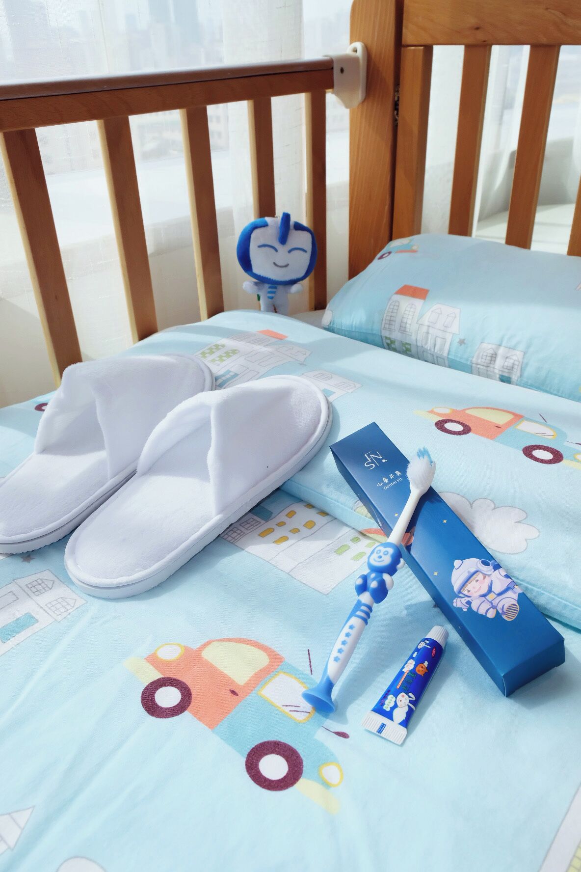 A child's bed with a blue mattress featuring cartoon cars and houses, white slippers, a toothbrush, and a toothpaste tube.