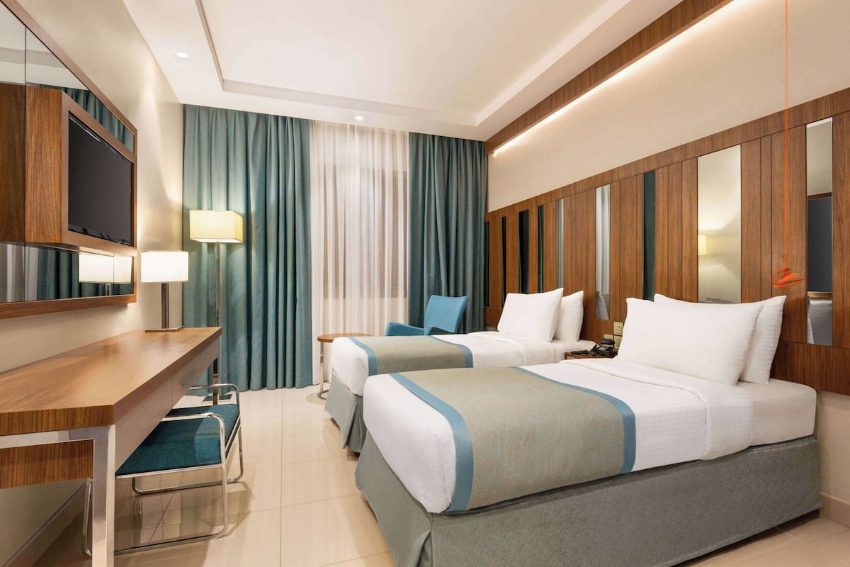 A hotel room with two beds, a desk, a chair, a TV, and a lamp.
