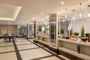 Un moderno vestíbulo de hotel con zona de comedor, mostrador de recepción y bufé con diversos platos y plantas.
