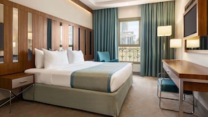 Ein Hotelzimmer mit einem großen Bett, einem Schreibtisch, einem Sessel, einem Fernseher und einem Fenster mit Blick auf Gebäude.