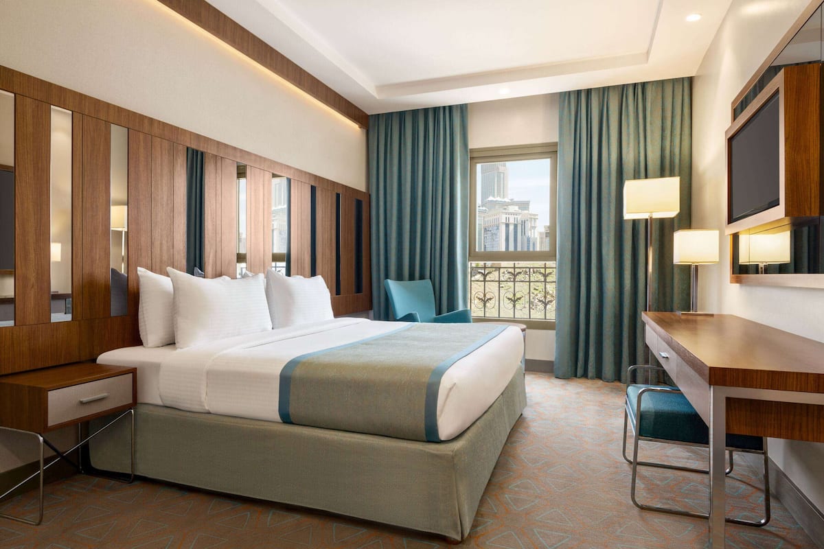 A hotel room with a large bed, a desk, a chair, a TV, and a window with a view of buildings.