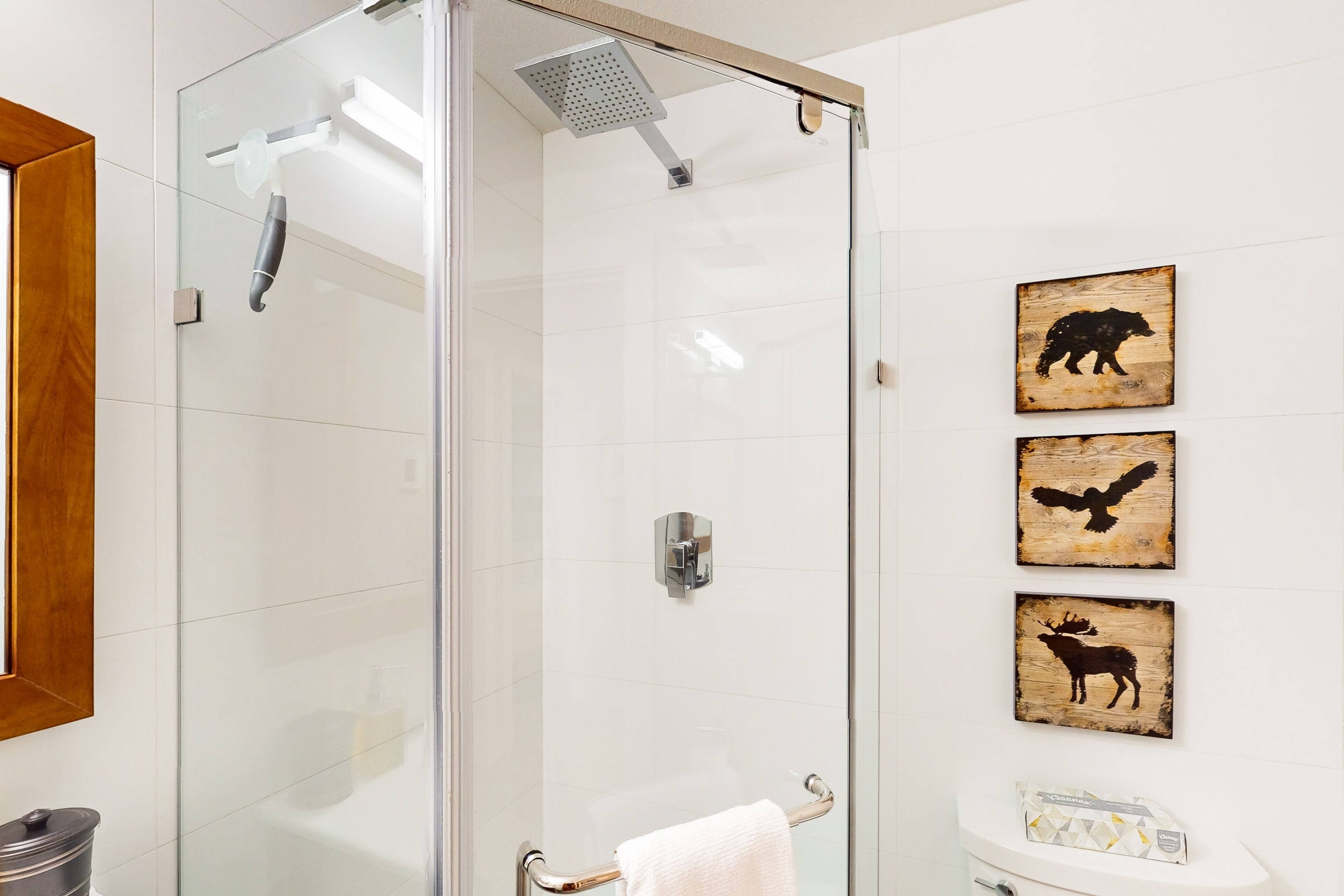 A glass-enclosed shower with a handheld showerhead, a towel rack with a white towel, and three wooden wall art pieces depicting a bear, an eagle, and a moose.
