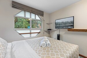 Un dormitorio con cama, ventana con vista a árboles, televisor en la pared y repisa con objetos.