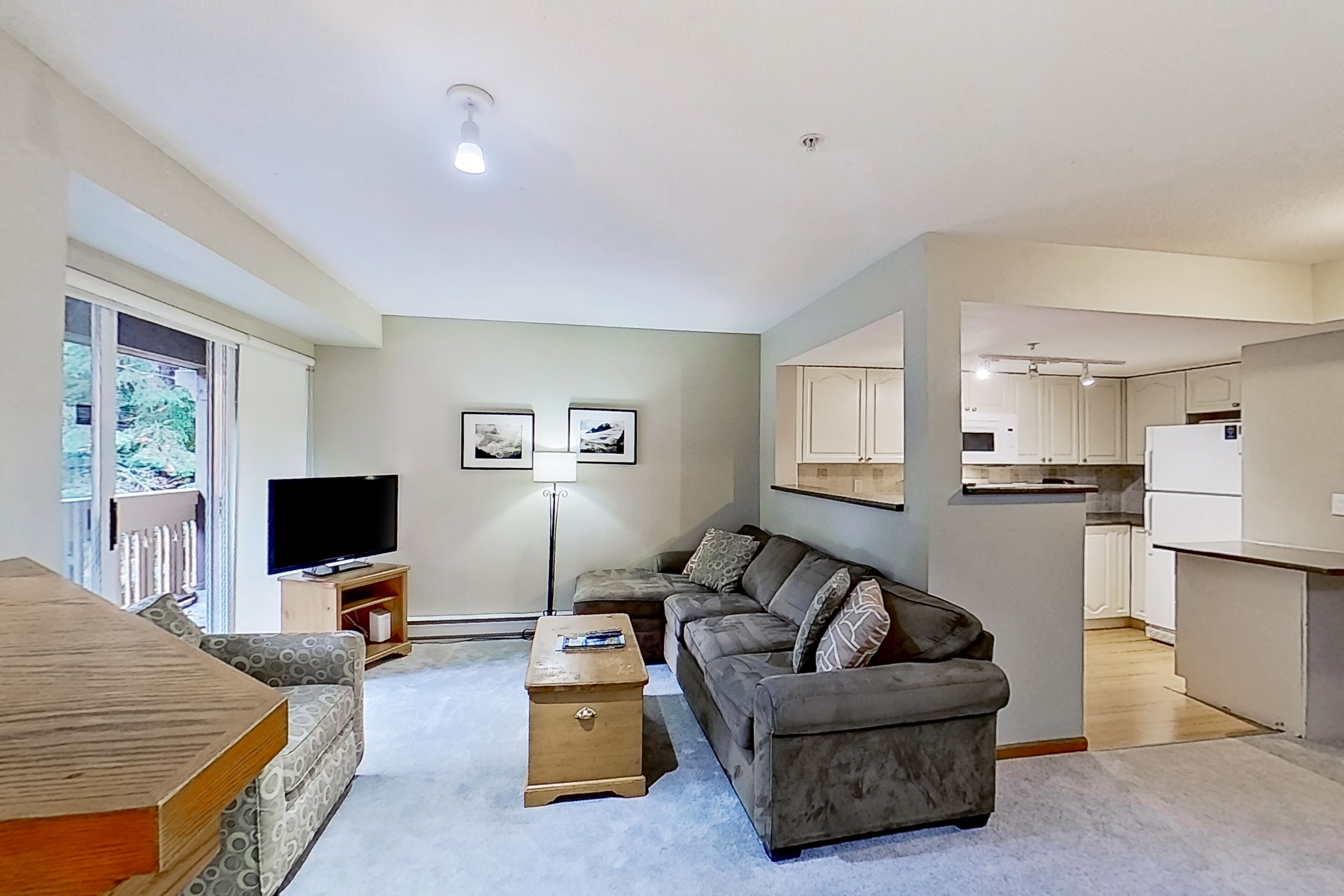 A living room with a sofa, a TV, a coffee table, and a dining table. There is a kitchen area visible in the background.