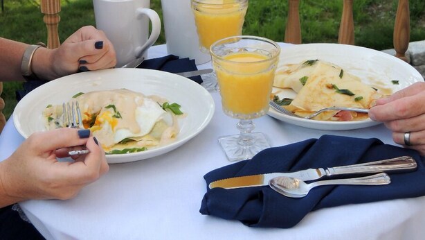 A dining table with plates of food, glasses of orange juice, and cutlery, set on a balcony with a view of a grassy area and trees. - Chandler River Inn (Jonesboro)