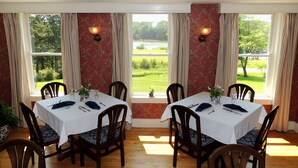 A dining room with a table set for four, large windows with curtains, and a view of a green landscape. - Chandler River Inn (Jonesboro)