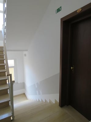 A staircase with a wooden handrail leading to a dark wooden door. A green exit sign is visible on the wall. - Hotel Marta Forlì (Forlì)