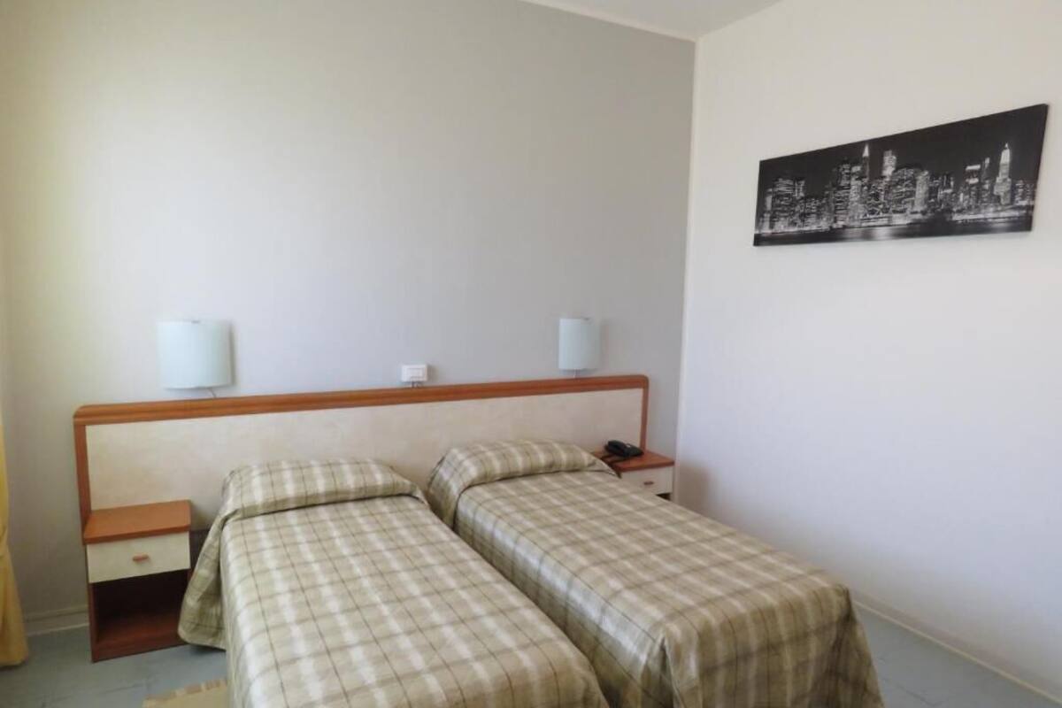 A bedroom with two beds, a nightstand, a lamp, and a cityscape painting on the wall.