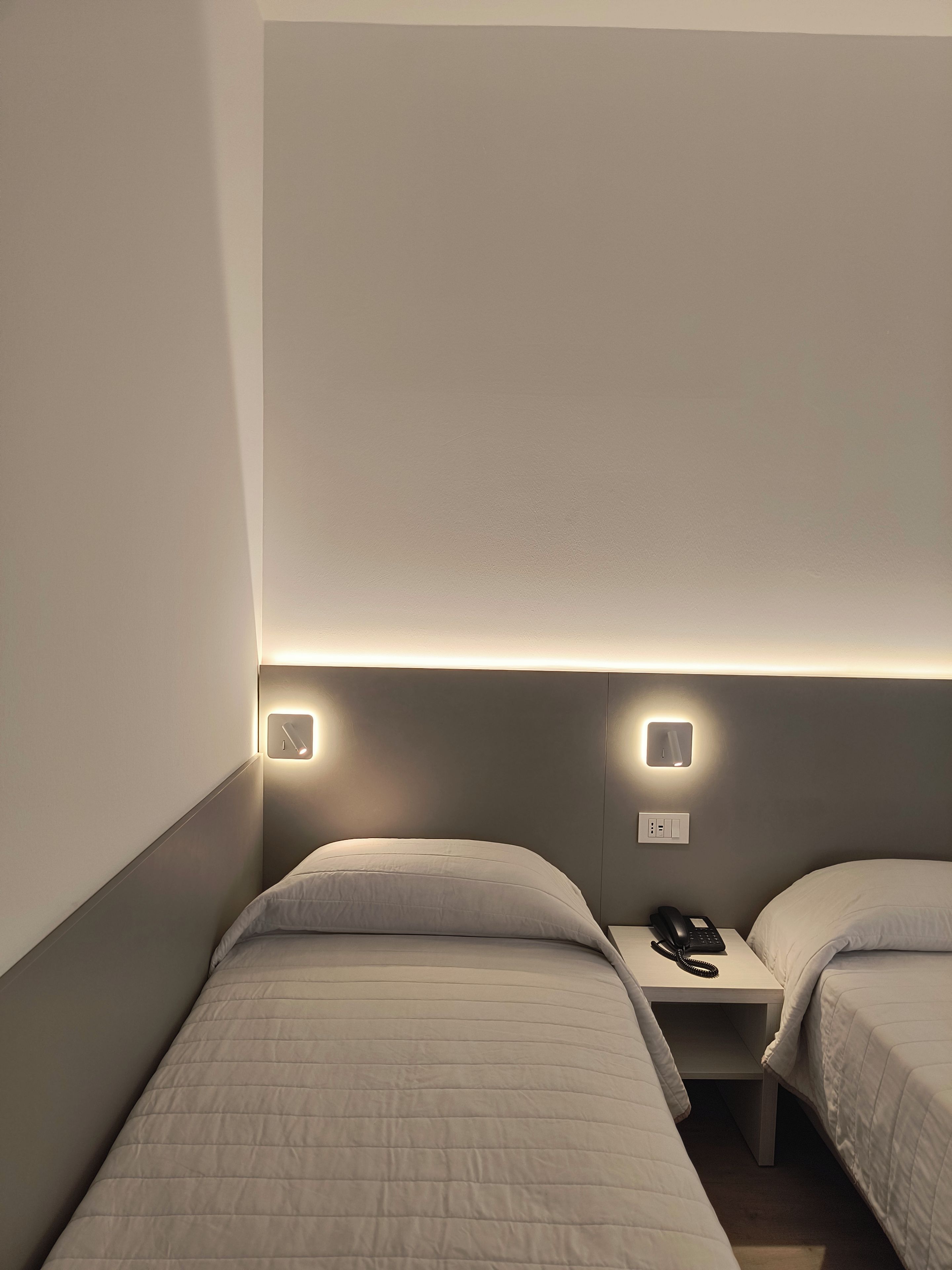 Una habitación de hotel con dos camas individuales, una mesita de noche con teléfono e iluminación en la pared.