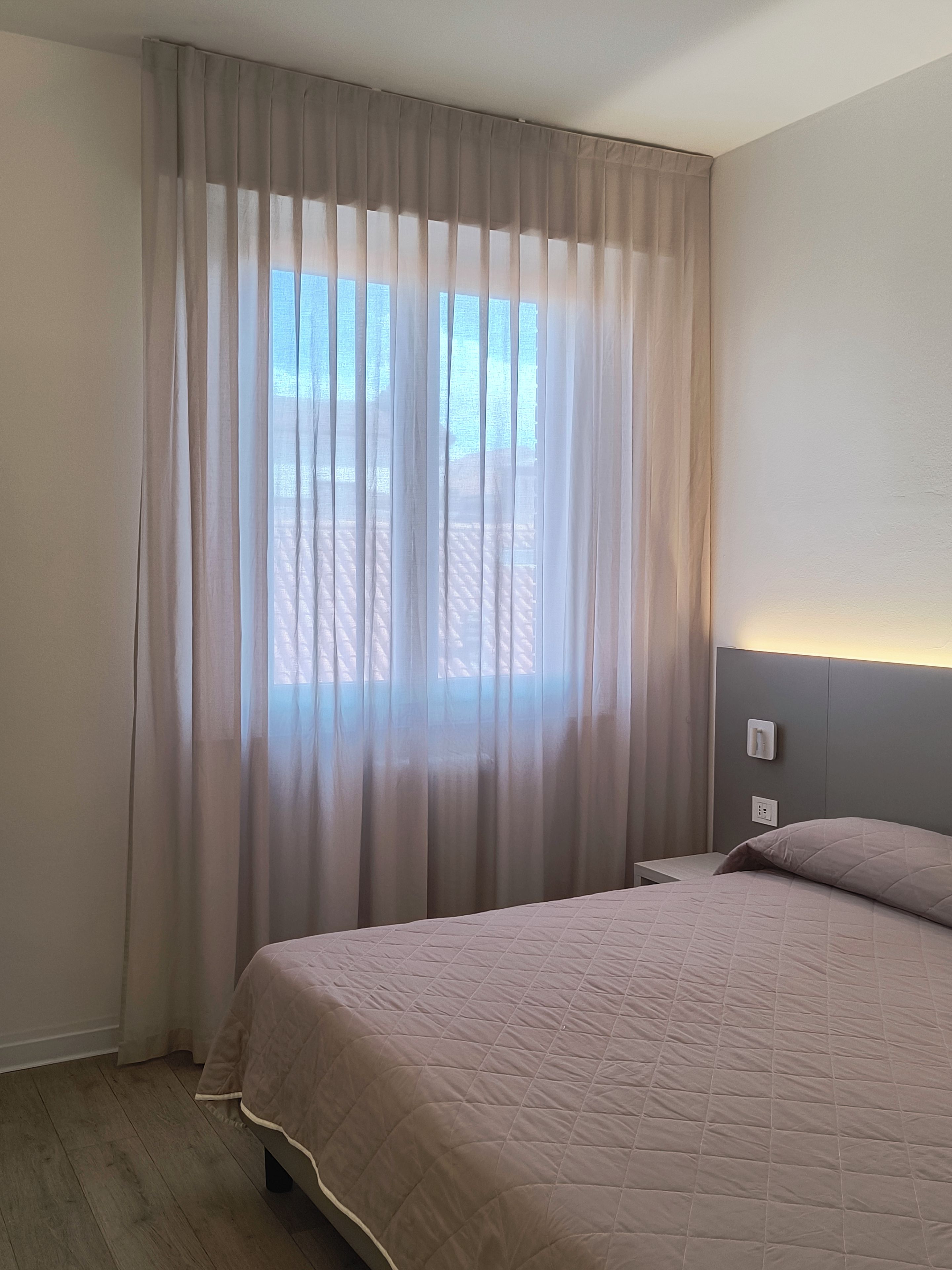 Un dormitorio con cama, cortinas, cabecera con luz y ventana con vistas al exterior.
