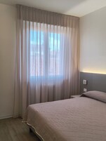 Un dormitorio con cama, cortinas, cabecera con luz y ventana con vistas al exterior.