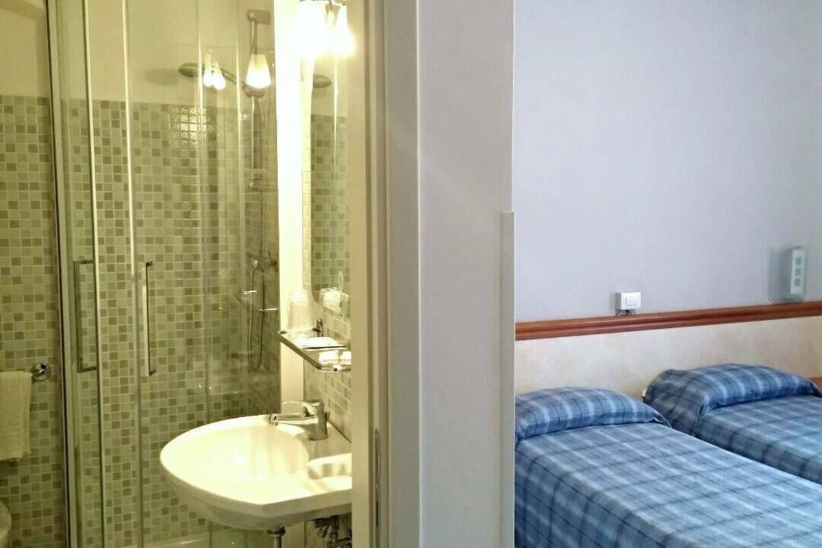 A small hotel room with a shower, a sink, and two beds with blue bedding.
