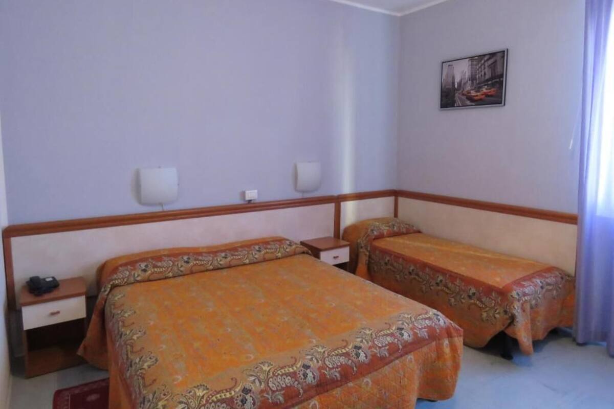 A hotel room with two beds, a nightstand, a wall-mounted lamp, and a painting on the wall.