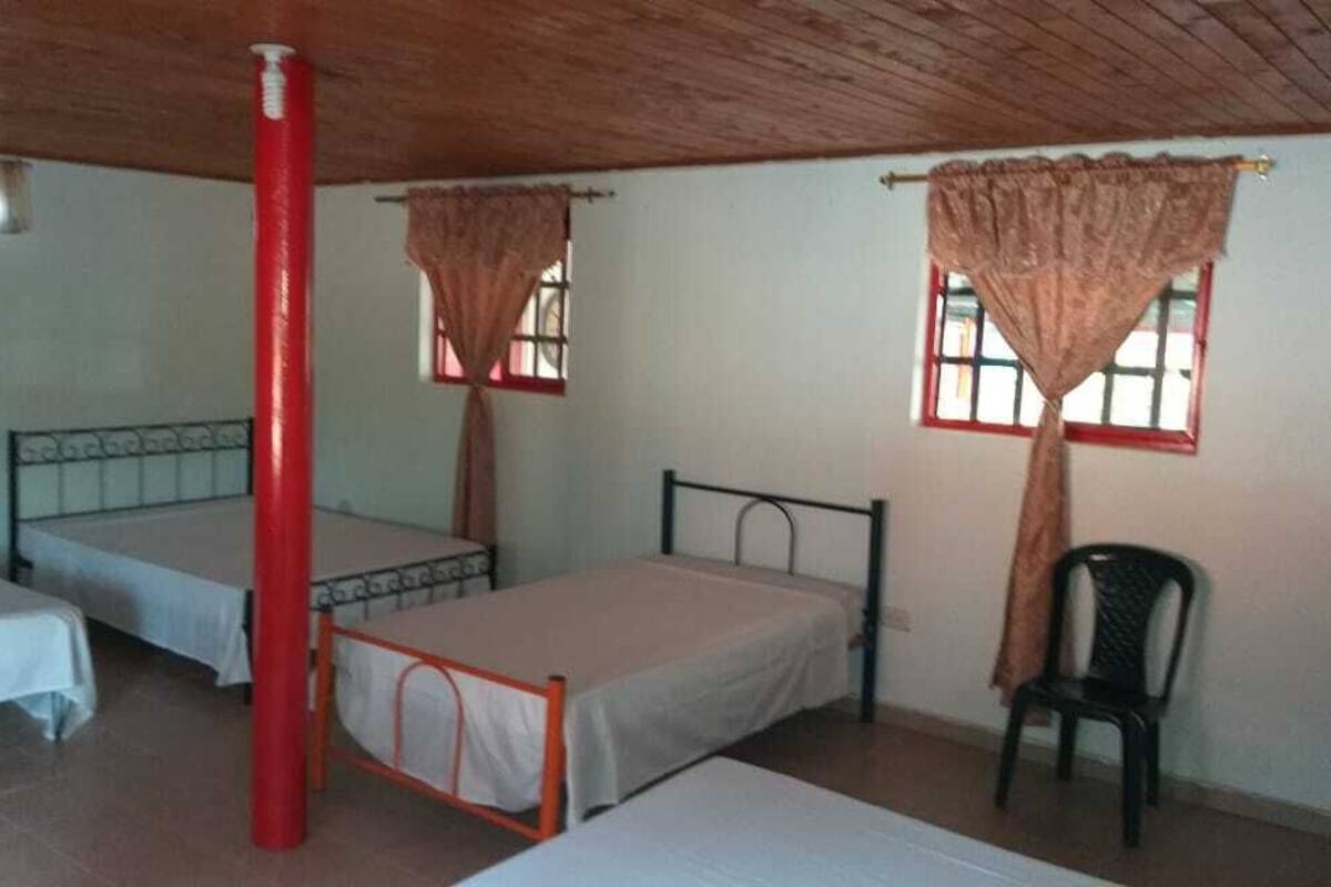 Una habitación con tres camas individuales, un pilar rojo y dos ventanas con cortinas.