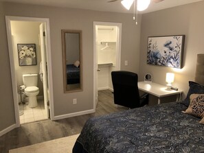 Un dormitorio con cama, escritorio, silla, espejo, lámpara de noche y un baño visible a través de una puerta abierta.