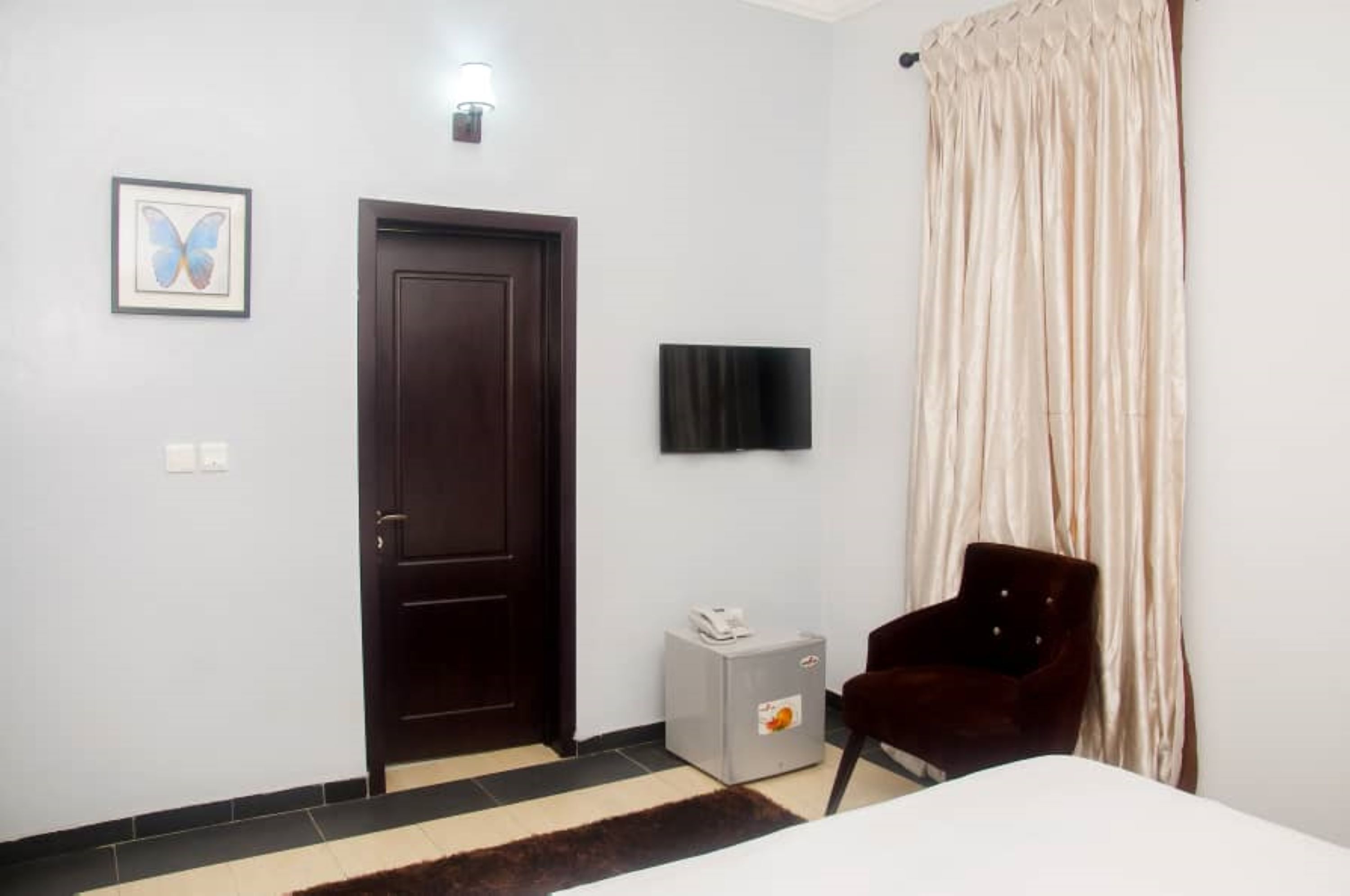 A hotel room with a dark wooden door, a flat-screen TV mounted on the wall, a small white cabinet with a phone, a dark red chair, and a large window with beige curtains.