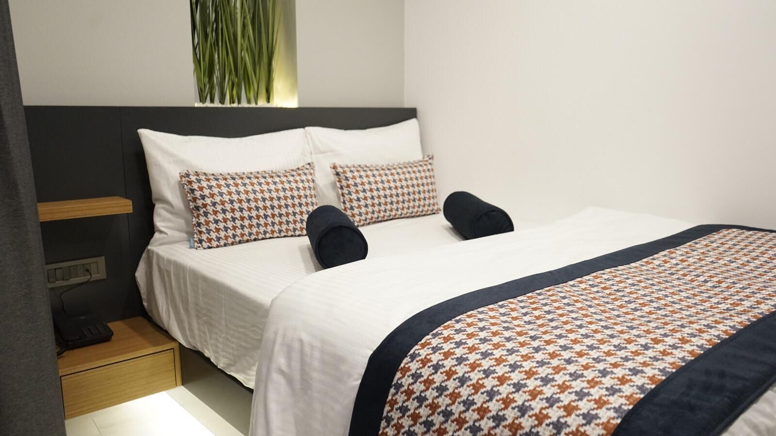 A neatly made bed with patterned pillows and a matching throw, a bedside table with a lamp, and a wall-mounted plant.