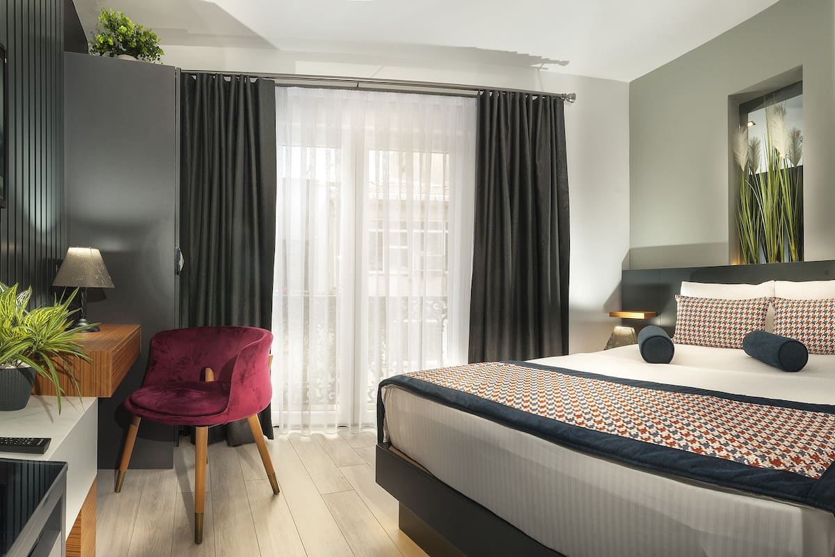 A modern hotel room with a large bed, a red chair, a desk with a lamp, and a TV on the wall.