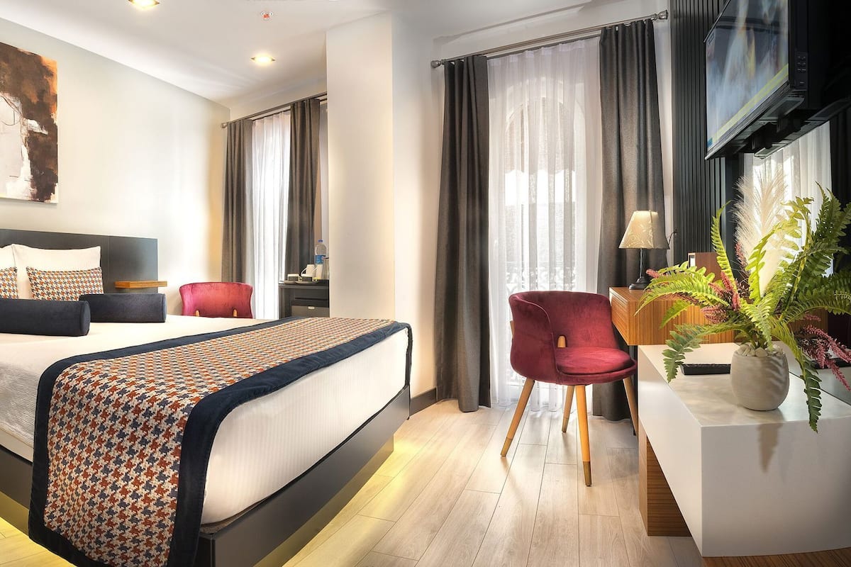 A modern hotel room with a large bed, a desk with a lamp, a red chair, and a large window with curtains.