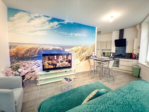 Ein Wohnzimmer mit Fernseher, einer Couch, einer Küchenzeile und einem großen Wandbild.