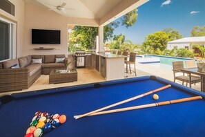 A pool area with a blue pool table, pool cues, and balls, a sofa, a television, and a bar area with outdoor seating.