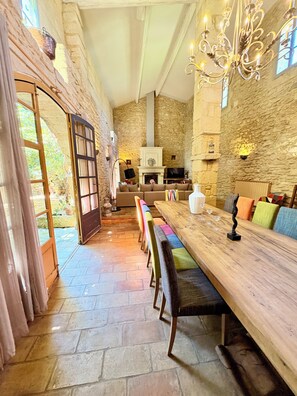 Een ruime kamer met een lange houten tafel, banken, een open haard, stenen muren en een kroonluchter.
