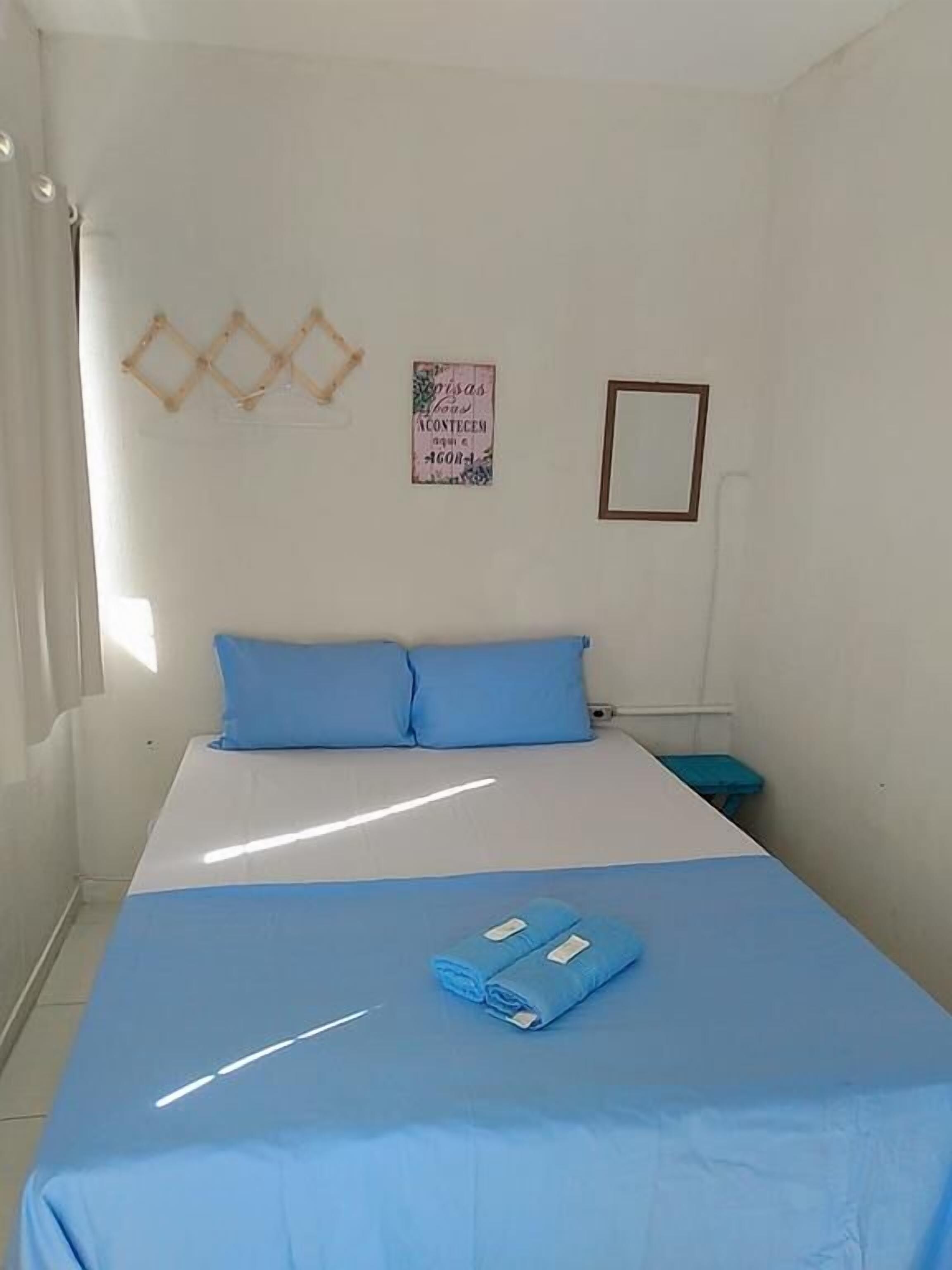 A bedroom with a bed, pillows, a blue blanket, a rolled-up towel, a small table, and a wall decoration.