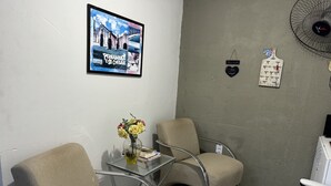 A waiting area with two beige chairs, a glass table with a vase of yellow flowers, a framed picture on the wall, and a wall-mounted fan. - Casa do Meio Pousada (Recife)
