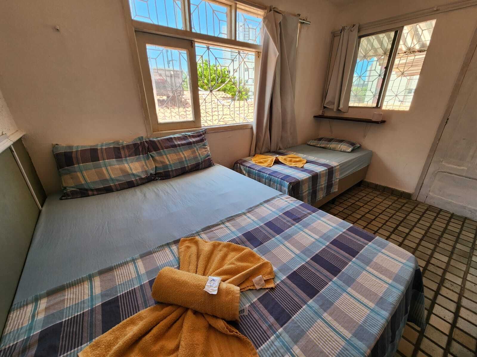 A room with two beds, each with a plaid blanket and a pillow. There is a window with curtains, and the room has a tiled floor.
