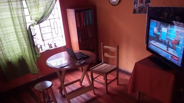 A room with a wooden floor, a small round table with a laptop, a wooden chair, a television on a stand, and a bookshelf. - Casa Refugio House (Cusco)